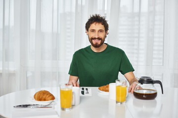 Fototapeta na wymiar cheerful attractive man in casual attire sitting at table during breakfast and smiling at camera