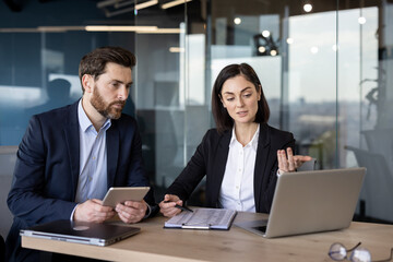 Successful business partners sitting by table with digital devices and looking at pc screen...