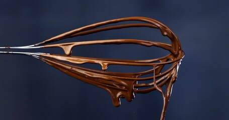 Super slow motion close up of liquid dark cocoa chocolate cream for bakery dessert preparation falling with splashing dripping drops from metal mixing whisk isolated on colorful background at 1000 fps