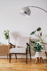 Stylish modern Scandinavian interior of living room with candles and fresh flowers on wooden console, retro style armchair, floor lamp and potted green home plants. Cozy home interior design. Vertical