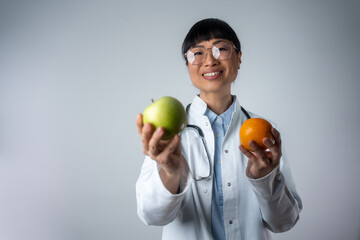 Smiling Asian dietician standing isolated and holding an orange and apple. Smiling Japanese woman doctor with a green apple and orange.