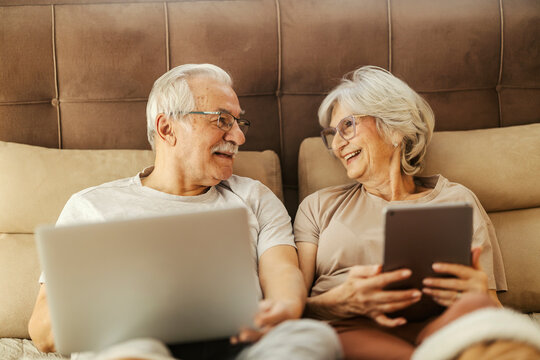 A happy senior couple is lying in a bed and using technologies.