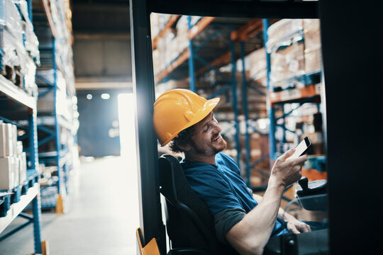 Male forklift operator in hard hat using smartphone in warehouse