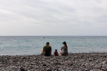 Family with small child sitting on idyllic volcanic black stone beach of Praia Garajai, Canico, Madeira island, Portugal, Europe. Waves hitting shoreline of majestic Atlantic Ocean. Vacation concept