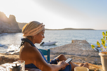 Relaxed beautiful woman checking her mobile phone while having breakfast in a typical Greek tavern...