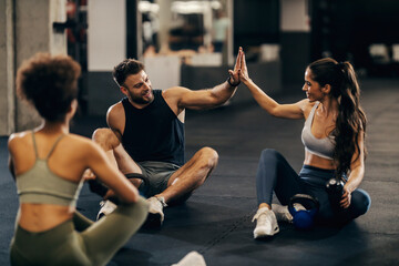 Group of three fit friends giving high five after exercise while sitting on a gym floor.