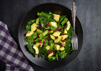 Avocado salad with spinach and pomegranate in a black plate on black background.