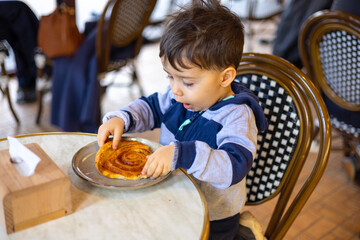 baby boy in pastry eating puff snail sitting at table on chair. smiling kid child with happy...