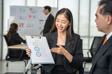 Cheerful Asian businesswoman presenting a clipboard with ESG (Environmental, Social, and Governance) concepts to a male colleague.