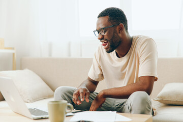 Smiling African American Man Working on Laptop at Home Sofa Happy young African American freelancer...