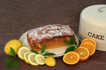 Lemon and orange lemon drizzle icing cake with fresh fruit and leaves and cake tin on rustic wood background. Homemade dessert for afternoon tea.