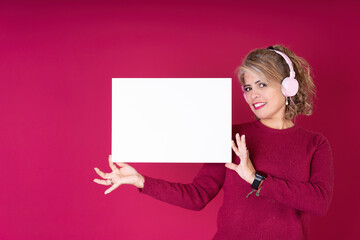 Portrait of a cheerful pretty latin woman wearing pink headphones holding and showing a blank empty square sign isolated on red studio background