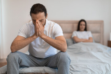 Stressed man sitting on bed with hands on face, woman in background