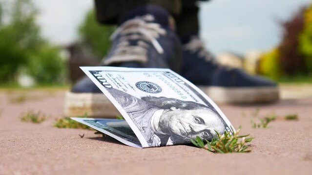 Close-up of a hundred dollar bill lost on the sidewalk and a walking man picks it up