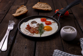 Fried eggs with bacon and vegetables set in cast iron frying pan, on old dark wooden table background