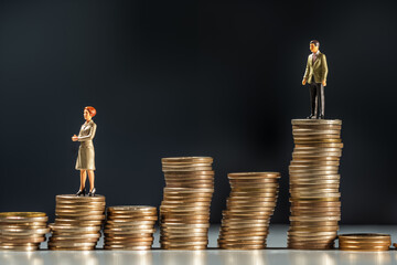 Miniature people: Businesswoman and businessman standing on a stack of coins on black background