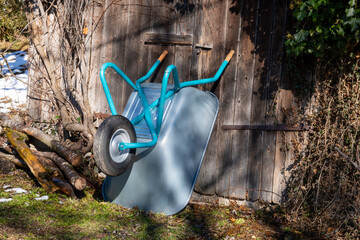 Wheelbarrow with a petrol-colored frame leans against the wooden wall of a shed in a rural area.