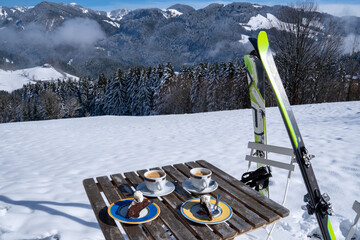 Break from skiing. The table is set with coffee and cake, the skis and ski poles are leaning against the chair. View of Chiemgau Alps and Geigelstein (1808 m), Bavaria, Germany in winter. - obrazy, fototapety, plakaty