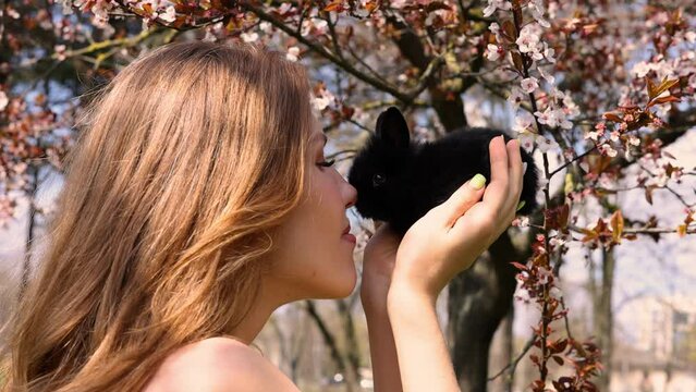 woman and black cute little bunny touching noses against spring blossom tree branches.girl and blonde girl nose to nose with rabbit.easter is coming, happy smiling female