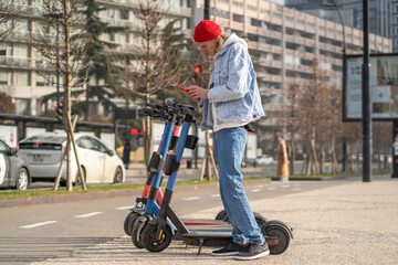 Focused hipster guy using mobile application in smartphone to rent electric scooter for driving in...