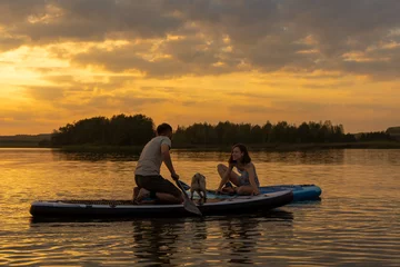 Foto op Canvas Happy couple paddle boarding at lake during sunset together with pug dog. Concept of active family tourism and supping with pets. Brave Dog Standing on SUP Board and enjoy lifestyle on summer vacation © Yekatseryna