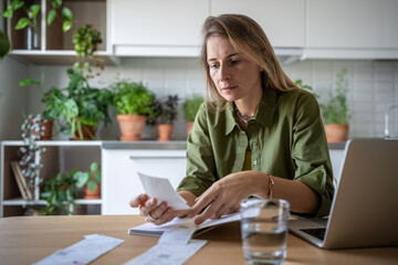 Thoughtful female sitting at table, holding bills, receipts, checking figures attentively. Serious woman working as freelance accountant at home doing financial analysis of company expenses and income