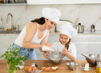 Mom and little girl prepare dough for baking, mom sifts and adds flour to bowl, girl mixes...