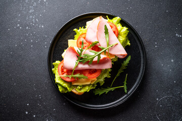 Ciabatta sandwich with lettuce, cheese, tomatoes and ham on black table.