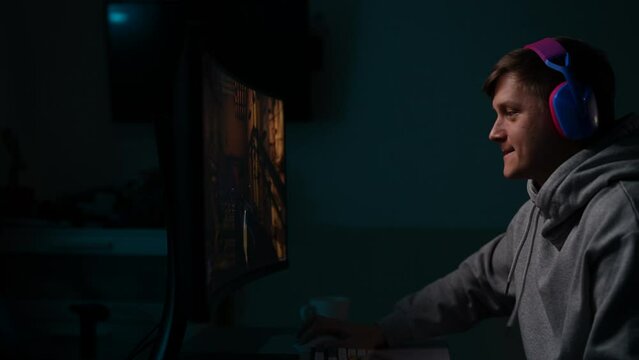 Tracking shot of happy gamer male in headphones playing first-person shooter online video game on powerful personal computer sitting at desk in dark room. Concept of modern online entertainment.