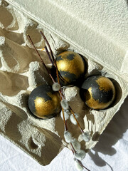 Black and gold eggs lie in a tray on a light background under the rays of the sun, next to a bunch of willow. Celebration of the bright Easter holiday. Vertical photo