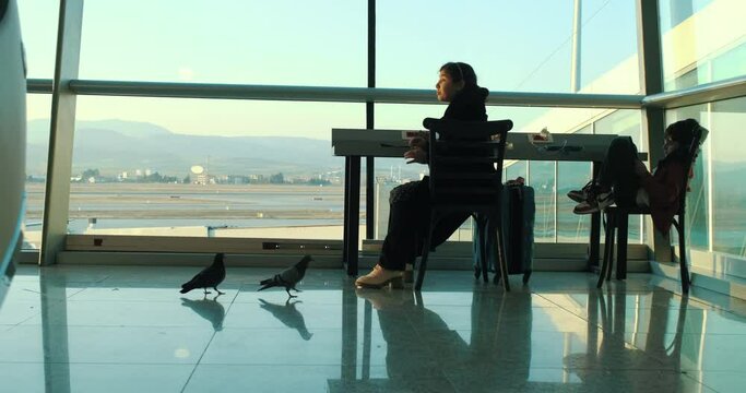 Pigeons eat at the airport terminal. Two children sitting in the terminal. One of them feeds the birds. The other one is playing with the tablet.