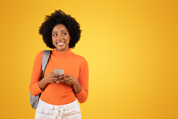 Happy young woman with afro hair, dressed in an orange turtleneck, texting on her smartphone