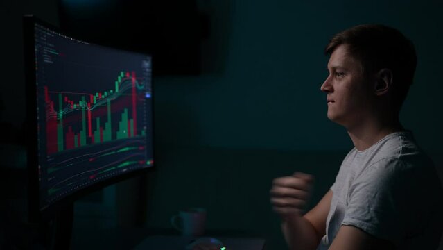 Happy excited male trader pounding fist on chest encouragingly in time with movements on stock market chart, sitting in front of computer monitor, looking at screen in dark room, slow motion.