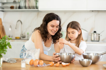 Obraz na płótnie Canvas Mom teaches her daughter how to cook dessert, shows how to beat dough with whisk. Mom and girl are talking in kitchen and mixing ingredients for pastry in bowl with whisk