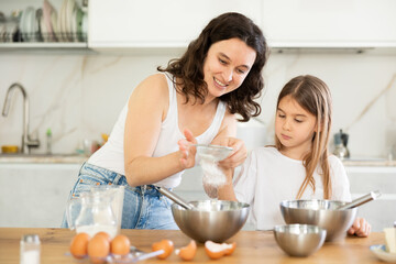 Obraz na płótnie Canvas Mom and little girl prepare dough for baking, mom sifts and adds flour to bowl. Family activity, concept of happy family and parenting. Homemade food and little helper.