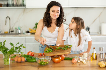 In kitchen, mom and daughter are cutting fresh vegetables to make salad. Groom explains to girl...