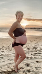 Fototapeta na wymiar Eine Junge schwangere Frau steht am Meeresstrand in einem schwarzen Bikini und streicht lächelnd über ihren Bauch. KI generiert.