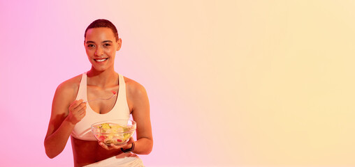 Healthy lifestyle concept with a beaming woman in sports attire, holding a bowl of fresh fruit salad