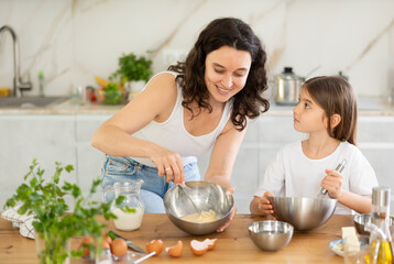 In kitchen, while preparing dough, mom teaches her daughter how to prepare dough for dessert, tells step-by-step recipe and shows how to work with whisk to get successful dough