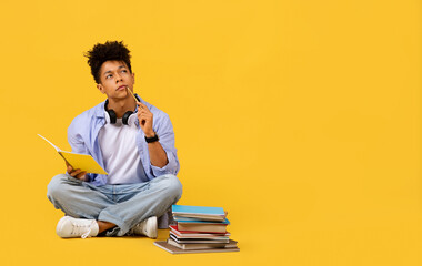 Thoughtful black male student with notebook and headphones on yellow background