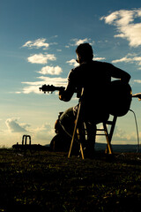 Silhueta de cantor tocando violão de frente com o por-do-sol e céu azul. 