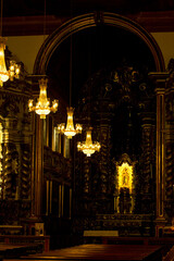 Interior de igreja católica com imagens de santos, e sequência de lustres acesos pendurados no teto.  