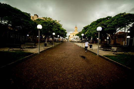 Pra&ccedil;a da cidade de Itu com igreja matriz de cor amarela, ruas, &aacute;rvore, poste de rua com luz acesa e c&eacute;u com nuvens carregadas. 