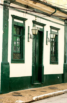 Fachada de casa com parede branca, janela de cor verde, porta de madeira, Arandela e calçada de pedra. 