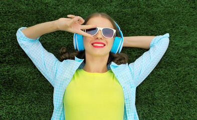 Happy smiling young woman listening to music in headphones while lying on grass in summer park