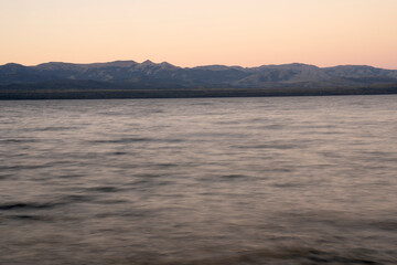 Long exposure shot of Nahuel Huapi lake and the Andes mountains in the background, at sunset. Beautiful blurred water effect, the rocky shore and dusk colors in the sky.
