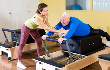 Female instructor of pilates helping elderly man workout. Two people working in pilates studio, woman assistant supporting and correcting old man patient beginner.