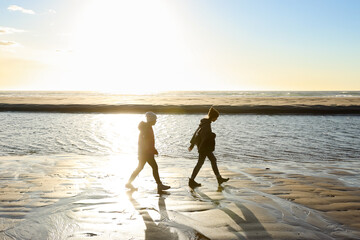 Fototapeta na wymiar Teen girl and tween girl walking along the coastline on a sandy beach at sunset, with the ocean and horizon behind them.