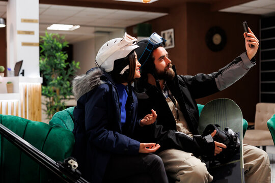 Side-view shot of active couple wearing winter clothing taking a selfie with mobile phone at lounge area. Boyfriend holding smartphone for picture with girlfriend at ski resort wintersport activity.