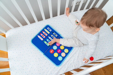 Baby playing with montessori busy book sitting in crib. Educational books and quiet books concept. Montessori busy board